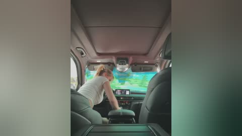 Media: Video of a woman with light skin and a ponytail, wearing a white tank top, driving a car with a green screen in the background.