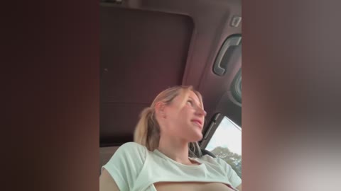 Media: Video of a blonde woman with a ponytail, wearing a white T-shirt, seen from behind while driving. The interior of the car is visible, with a brown headrest and a rear-view mirror.