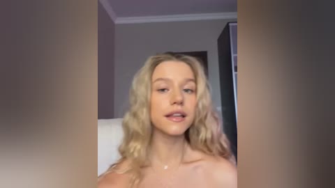 Media: Video of a topless, fair-skinned, blonde woman with wavy hair, looking seductively into the camera, set in a dimly lit, modern room with muted colors and a white ceiling.