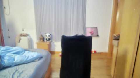 Media: Video of a bedroom with a black chair in the foreground, a bed with blue sheets, a panda plush toy on a wooden nightstand, and white curtains covering a window.