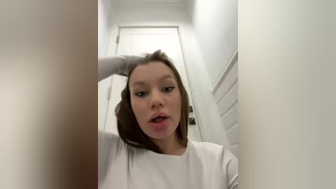 Media: Video of a young woman with fair skin and straight brown hair, wearing a white top, captured from a low angle in a narrow hallway with white walls and a closed door in the background.