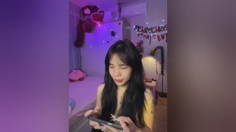 Media: Video of an Asian woman with long black hair, wearing a black top, seated in a dimly lit room with a \"Welcome Home\" banner, playing a video game.