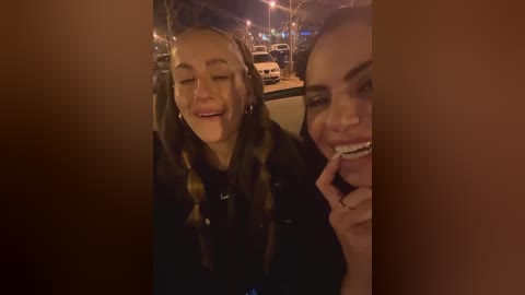 Media: Video of two young women with medium skin tones, smiling, in a dimly lit car. They have straight hair and wear dark coats. Background shows streetlights, parked cars, and bare trees.