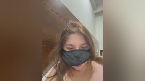 Media: Video of a young woman with long brown hair, wearing a black mask, indoors, possibly in a bathroom, with wooden paneling and a white ceiling.