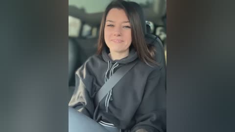 Media: Video of a smiling young woman with long, dark hair, wearing a black hoodie, seated in a car with a gray seatbelt across her chest.