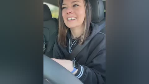 Video of a smiling, light-skinned young woman with shoulder-length brown hair, wearing a black hoodie and driving a car with gray seats.