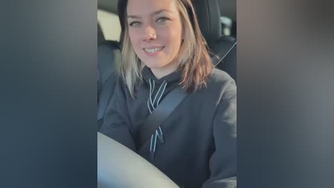 Video of a young woman with light skin, blonde hair, and green eyes, wearing a dark grey sweater and a striped tie, seated in a car with seat belts visible.