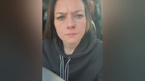 Media: Video of a young woman with fair skin and brown hair, wearing a black hoodie, looking slightly annoyed. Background is blurred, with a hint of an indistinct scene.