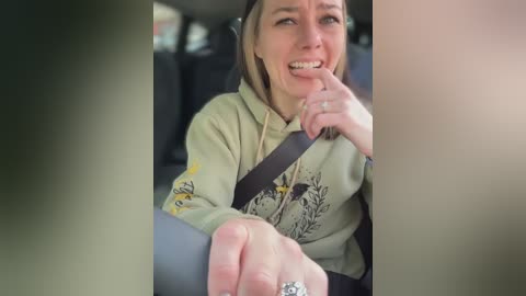 Video of a crying woman in a car, wearing a beige hoodie with a graphic design, seated in the passenger seat, holding a man's hand, and wearing a ring.