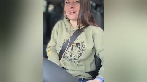 Video of a young woman with long brown hair, smiling, wearing a green hoodie with a graphic design, seated in a car with a shoulder strap.