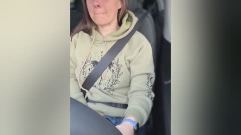 Media: Video of a woman in a car, wearing a green hoodie with a floral design, seated in the driver's seat with a seatbelt on, looking slightly to the side.