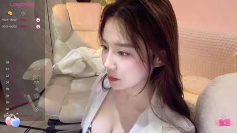 Media: A video of a young East Asian woman with fair skin, brown hair, and large breasts, wearing a white jacket over a black bra, sitting on a beige sofa.