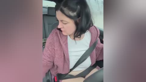 Media: Video of a woman with shoulder-length dark hair, wearing a white shirt and pink cardigan, buckled into a car seat with a seatbelt, in a grey car interior.