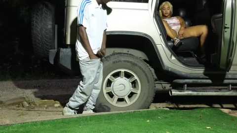 Media: Video: A young Black man in a white polo and gray sweatpants stands beside a Jeep. A blonde woman in a tank top and shorts sits in the open Jeep door, looking distressed.