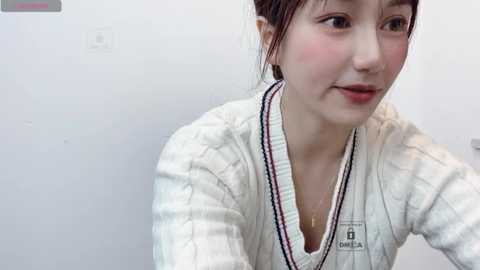 Media: Video of a young Asian woman with fair skin, brown hair, and red lipstick, wearing a white, ribbed knit sweater. She looks slightly to the right, with a neutral expression.