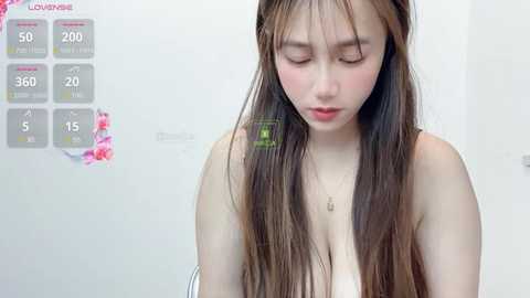 Media: A video of an East Asian woman with long brown hair, fair skin, and a slender build, wearing a white top, standing in a bathroom with a flowered wall calendar and temperature gauge.