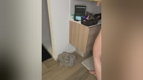 Media: A video of a partially nude woman standing in a room with a wooden floor, light-colored walls, and a wooden cabinet. A gray towel is on the floor near her feet.