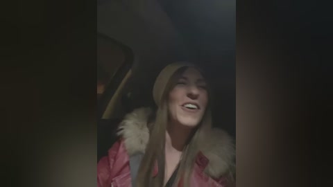 Media: A video captures a young woman with long blonde hair, wearing a pink fur-trimmed coat and a beige beanie, smiling joyfully in a dimly lit car interior.