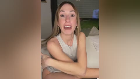 Media: Video of a young, fair-skinned woman with long blonde hair, wearing a white sleeveless top, sitting on a beige couch in a modern living room.