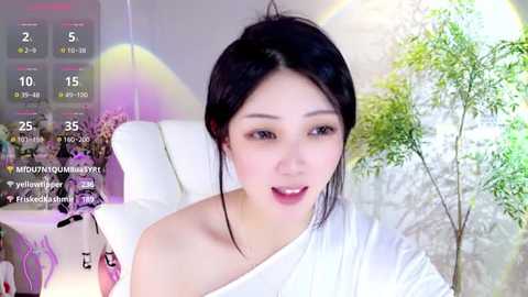 Media: A video of an East Asian woman with light skin, black hair, and a white off-shoulder top, smiling in a bright room with a green plant and a white chair.