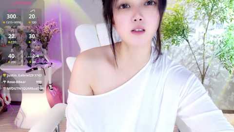 Media: A young Asian woman with fair skin and dark hair wears a white off-shoulder top, sitting in a bright, modern room with a white chair and plant.