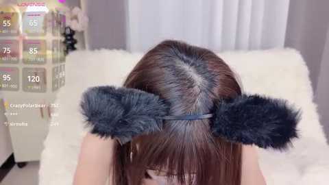 Media: A video showing a woman with straight, shoulder-length brown hair, wearing a dark grey, fluffy bunny ear headband in a cozy, light-colored room with a white furry blanket and a pink flower arrangement.