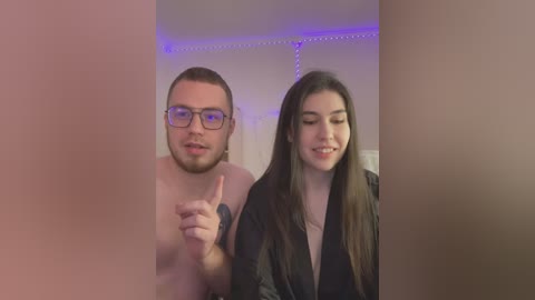 Media: Video of a bearded man with glasses and a woman with long hair, both smiling and gesturing towards the camera in a dimly-lit room with purple LED strip lighting.