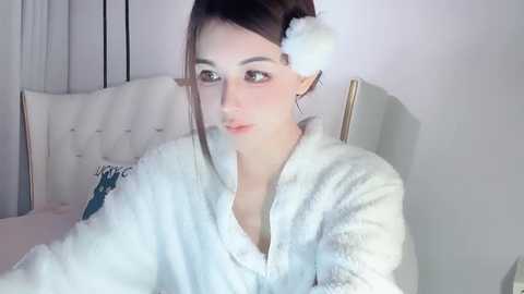 Media: A video of a young woman with fair skin and dark hair, wearing a fluffy white bathrobe, seated on a white tufted chair in a softly lit bedroom.