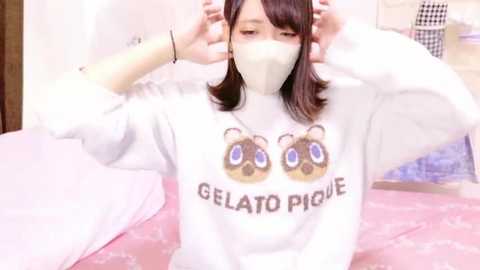 Media: Video of a young woman with light skin and shoulder-length brown hair, wearing a white sweatshirt with \"Gelato Pique\" and bear logo, a white mask, and adjusting her hair in a pink room.