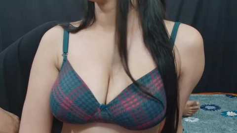 Media: Video of a light-skinned woman with long black hair, wearing a blue and green plaid bra with thin straps. She is seated, with a dark background and a patterned blue and white fabric visible behind her.