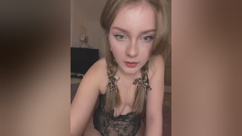 Video of a young woman with fair skin, light brown hair in braids, wearing a black lace bra, looking down. Background includes a TV and beige walls.