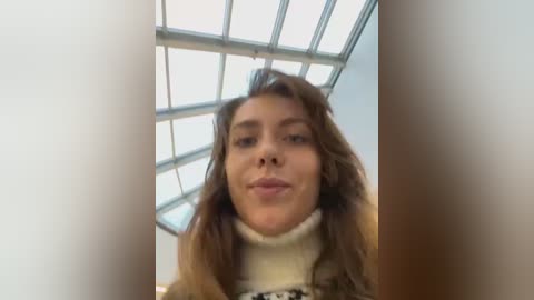 Media: Video of a young woman with wavy brown hair and light skin, wearing a cream turtleneck sweater, standing in a modern indoor space with a glass ceiling.