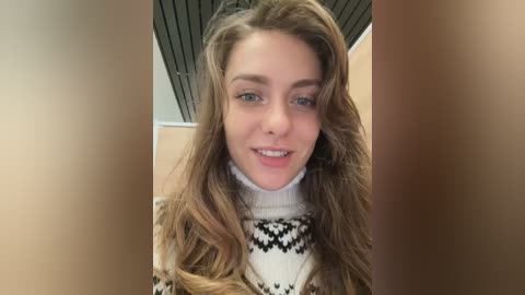 Media: Video of a young Caucasian woman with long, wavy, light brown hair, smiling, wearing a white turtleneck sweater with black geometric patterns. Background shows a ceiling with slatted wood panels and beige walls.