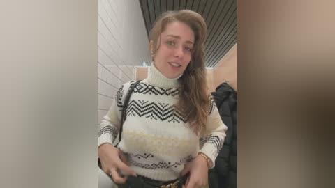 Media: Video of a young woman with light skin and long, wavy brown hair wearing a white sweater with black zigzag patterns and a yellow logo. She stands in a hallway with white tiles, black ceiling panels, and black coats hanging on a rack.