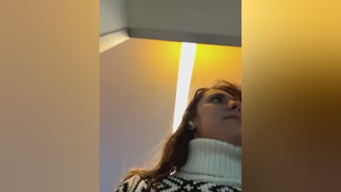 Media: Video of a woman with long brown hair, wearing a white turtleneck sweater, looking upward, with warm lighting casting a glow on a beige wall and ceiling.