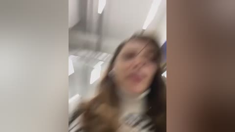 Media: Video of a young woman with long, light brown hair, wearing a white top, blurred due to motion, in a modern, bright indoor setting with white walls and fluorescent lights.