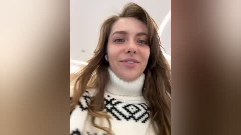 Media: Video of a young Caucasian woman with long, wavy brown hair, wearing a cream-colored turtleneck sweater with black geometric patterns. Background is blurred, with soft lighting.