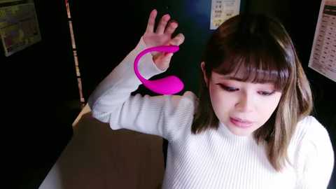 Media: Video of a young Asian woman with straight brown hair, wearing a white ribbed sweater, holding a pink vibrator, indoors with dim lighting and black walls.