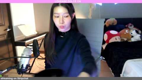 Media: Video of an Asian woman with long black hair, wearing a black turtleneck, seated at a desk with a computer and microphone, surrounded by plush toys.