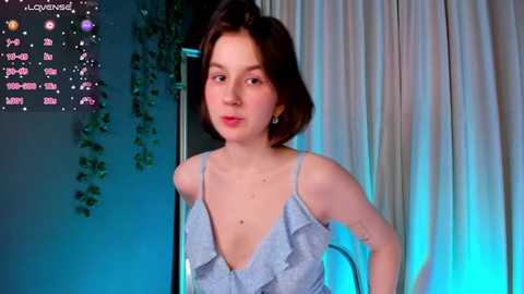 Media: Video of a young woman with short brown hair, wearing a low-cut, light blue ruffled top, standing in a dimly lit room with a calendar and greenery in the background.