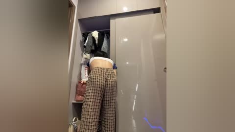 Media: A video of a person with light skin and a slender physique bending over to pick up items from a beige wardrobe. They wear loose, brown and white checkered pants and a white shirt. The wardrobe's interior is filled with various clothes, including a black jacket.