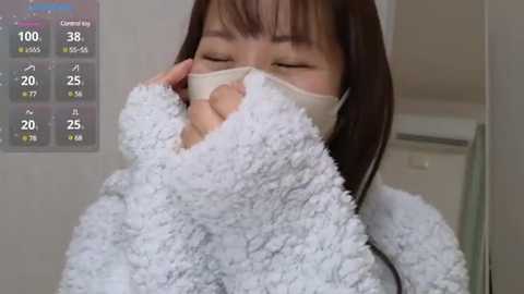 Media: A video of an Asian woman with straight, shoulder-length brown hair, wearing a white, fluffy bathrobe, holding a beige face mask, eyes closed, in a bathroom with a digital thermometer display.