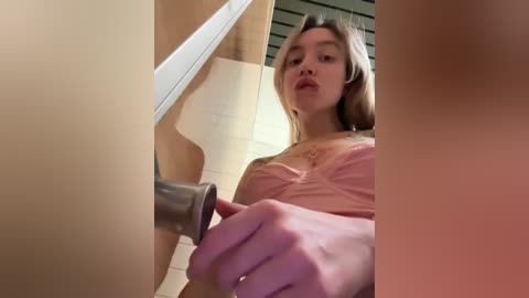 Media: Video of a young woman with light skin, long brown hair, and a tattoo on her chest, wearing a pink bra, inserting a metal dildo into a wooden door.