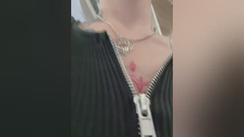 Media: Video of a woman with fair skin, wearing a black sweater with a silver zipper partially open, revealing a small red heart tattoo on her chest.