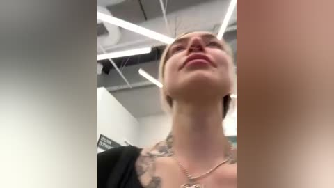 Media: A video of a smiling, tattooed woman with light skin, wearing a black top, in a brightly lit, modern room with white walls and exposed ductwork.