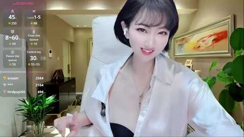 Media: Video of an East Asian woman with short black hair and light skin, wearing a white satin blouse, sitting at a desk. Modern office background with plants and framed artwork.