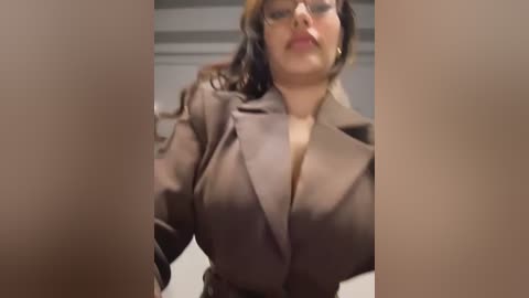 A video of a plus-size woman with dark hair and glasses, wearing a brown blazer, standing in a dimly lit room.