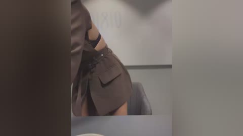 A video of a person in a brown suit, leaning forward, with a blurred background of a whiteboard and a chair. The image is cropped and out of focus.