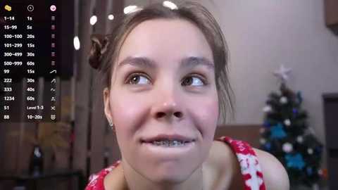 Media: Video of a young woman with fair skin and braces, wearing a red dress with white patterns, smiling against a blurred indoor background with a Christmas tree.