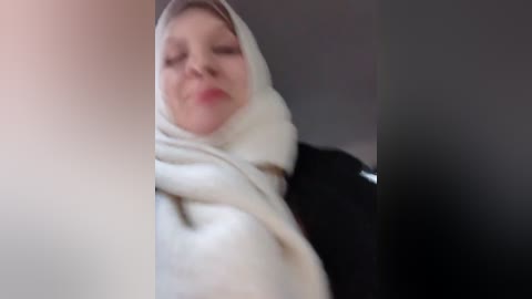 Media: Video of a woman with fair skin, wearing a white hijab and black coat, with a neutral expression, in a dimly lit room.